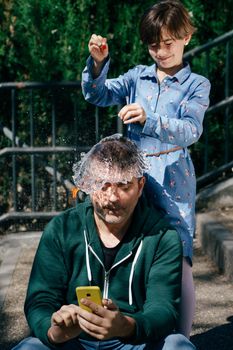 Little girl blowing up a water-filled balloon over her father's head to get him to stop staring at his smartphone.