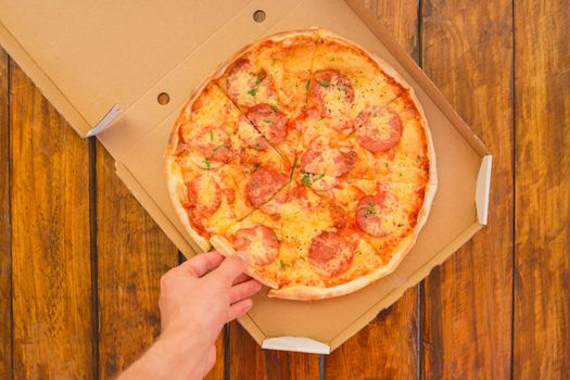 The guy's hand takes a pizza in cardboard box of against the background of a wooden table. Delicious fast food.