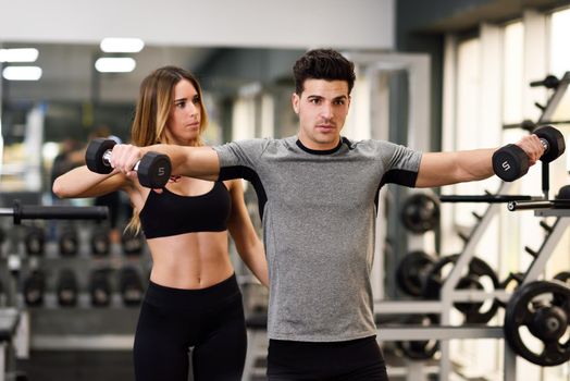 Female personal trainer helping a young man lift dumbells while working out in a gym