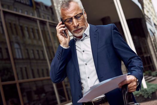 Senior businessman is discussing on the phone a contract outdoors near office