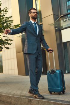 Catching taxi. Full length of young and handsome bearded man in suit carrying suitcase and raising his arm while standing outdoors. Travelling. Business trip