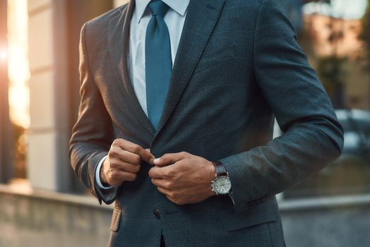 Cropped photo of a man adjusting his suit jacket while standing outdoors. Business concept. Stylish people