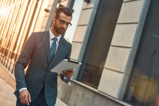 Working ewerywhere. Young and handsome bearded businessman in eyeglasses looking at his digital tablet while walking outdoors. Digital concept. Business concept