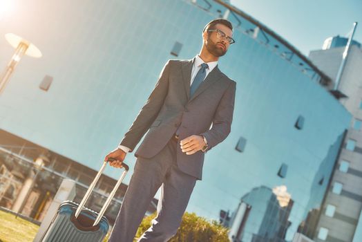 Business trip. Young and handsome bearded man in full suit pulling his luggage and looking away while walking outdoors. Business travel