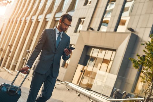 Always available. Young and handsome bearded man in suit pulling suitcase and looking at his smartphone while walking outdoors. Travelling. Business concept