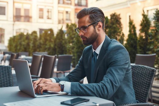 Modern businessman. Portrait of handsome bearded businessman in eyeglasses working with laptop while sitting in restaurant outdoors. Digital concept. Business concept