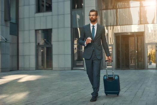 Busy morning. Full length of young and handsome bearded businessman in suit pulling his luggage and holding cup of coffee while walking outdoors. Travelling. Business concept