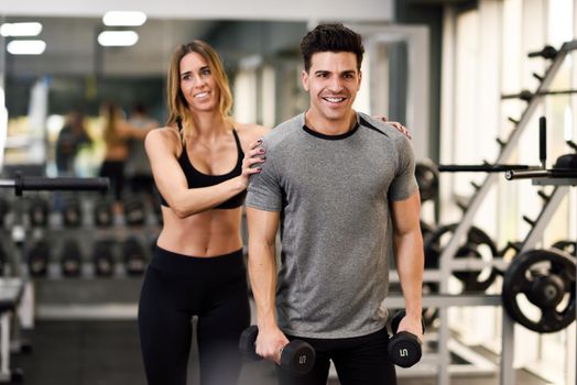 Female personal trainer helping a young man lift dumbells while working out in a gym