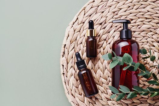 Dropper bottles of essential oil or serum, eucalyptus branches and dark dispenser bottle of shower gel on rattan background. Beauty and SPA concept. Flat lay, copy space