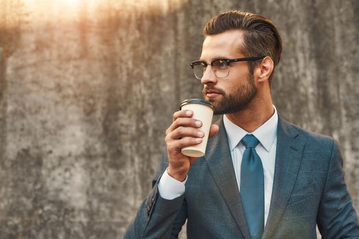 Confident and successful. Young businessman in formal wear and eyeglasses drinking coffee and looking away while standing against grey stone wall outdoors. Business concept