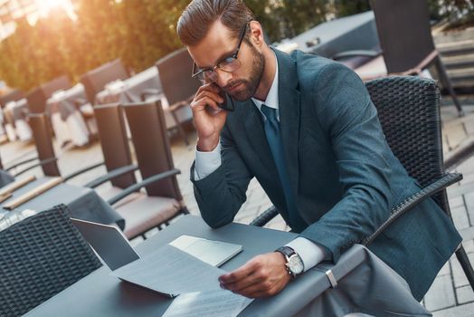 Working from everywhere. Portrait of handsome bearded businessman in eyeglasses talking by phone with client and looking at documents while sitting in restaurant outdoors. Digital concept. Business concept