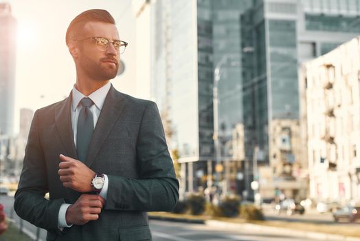 Portrait of young and handsome bearded man in full suit adjusting his sleeve and looking away while standing outdoors with cityscape in the background. Business concept