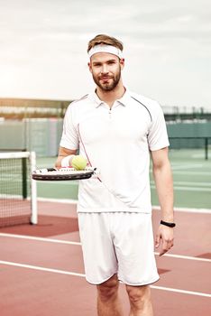 Portrait of handsome tennis player in white sportswear with the racket and the ball in his hand standing on the tennis court. Vertical shot