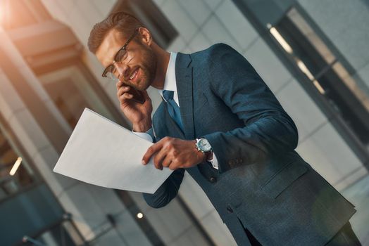 I have great new for you Portrait of smiling bearded businessman in full suit talking by phone with client and looking at documents while standing outdoors. Business concept