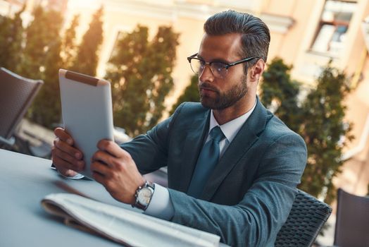 Modern businessman. Portrait of handsome bearded businessman in eyeglasses working with touchpad while sitting in restaurant outdoors. Digital concept. Business concept