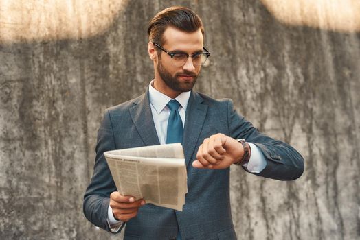 What time is it Young and handsome bearded businessman in eyeglasses checking the time and holding newspaper in one hand while standing against grey stone wall outdoors. Business concept. Time concept