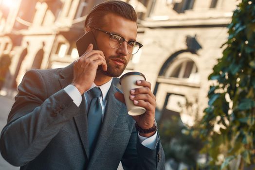 Important talk. Young and handsome bearded businessman in formal wear talking by phone with client and holding cup of coffee while standing outdoors. Business concept