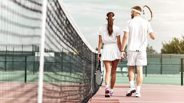 Young couple in white sportswear with the rackets in their hands is going to play tennis on the court