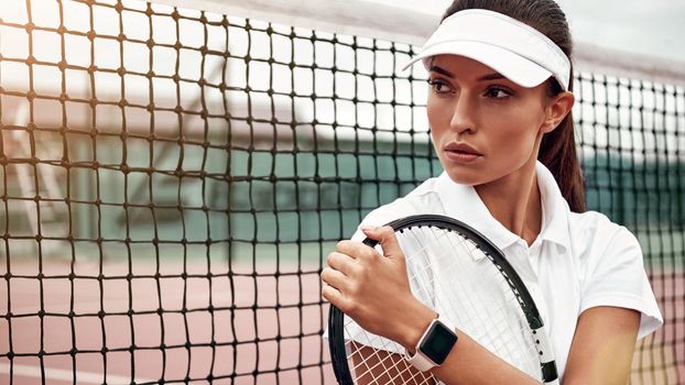Portrait of female tennis player sitting with a racket on a court, looking away. Healthy lifestyle. Horizontal shot