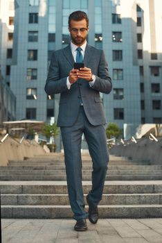 Business message. Full length of young and handsome businessman in formal wear using smart phone while walking outdoors. Business concept. Digital concept