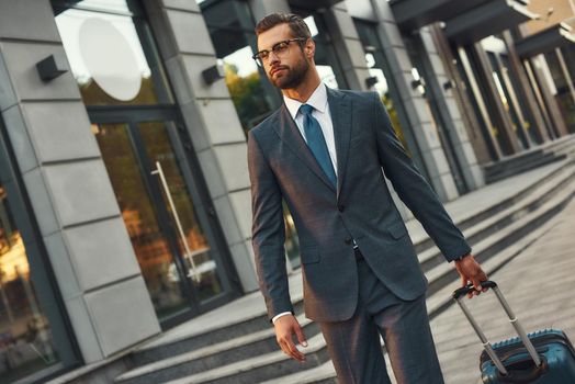 Business travel. Young and handsome bearded man in suit pulling his luggage and looking away while walking outdoors. Trip. Success