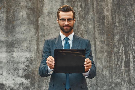 Modern businessman. Portrait of handsome bearded businessman in eyeglasses showing digital tablet at camera while standing against grey stone wall outdoors. Digital concept. Business concept