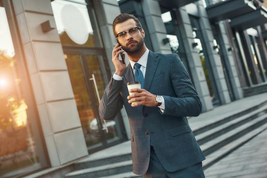 Important talk. Young and handsome bearded businessman in formal wear talking by phone and holding cup of coffee while standing outdoors. Business concept