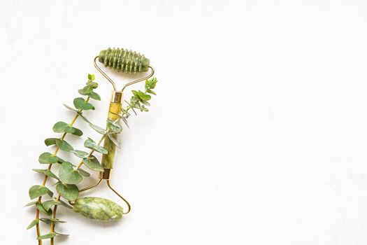 Green face roller from natural jade stone and eucalyptus branches on a white background. SPA concept. Top view, copy space for text