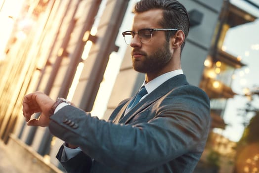 What time is it Young and handsome bearded businessman in eyeglasses checking the time while standing outdoors. Business concept. Time concept