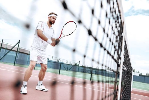 Handsome man holding tennis racket and looking concentrated while standing on tennis court