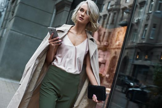 Fashion. Young stylish woman walking on the city street browsing internet on smartphone looking aside pensive