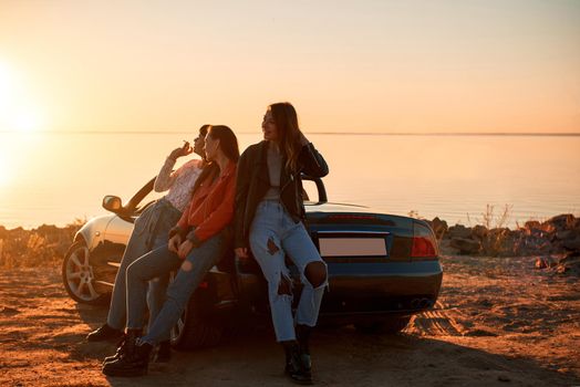 Cheerful young three women standing near cabriolet. Beautiful sunset at the background