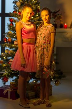 Beautiful Smiling Sisters Standing Together and Hugging in Front of Glass Wall near Christmas Tree, Two Adorable Teen Girls Wearing Nice Dresses Posing in Christmas Warm Cozy Room Interior.