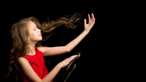 Inspired Beautiful Girl Wearing Red Nice Dress Sitting on Chair Posing at Camera, Portrait of Charming Girl with Flying Long Hair Looking to the Side Against Black Background