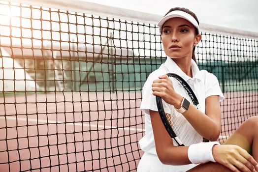 Portrait of female tennis player in white sportswear sitting with a racket on a court, looking away. Healthy lifestyle. Horizontal shot