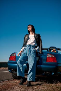 Young woman near roofless car and smiling outdoors on sunny day near the empty road. No people, sportive blue car