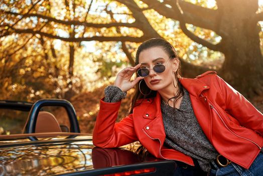 Beautiful girl near the car. Autumn season, red coat and jeans, stylish sunglasses on head