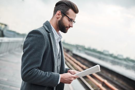 Business trip. Young businessman standing near railway reading newspaper close-up
