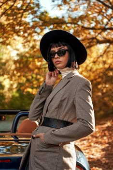 Attractive young woman posing near cabriolete. She wears black hat and sunglasses. Stylish autumn.