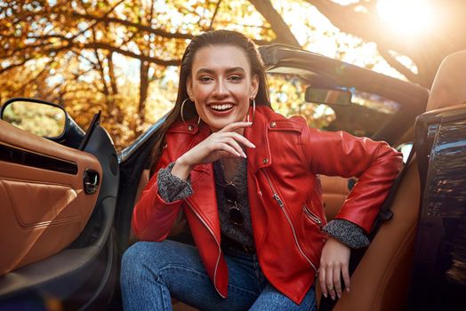 Beautiful girl in the roofless car. Autumn season, red coat and jeans, stylish sunglasses on head. Skin salon