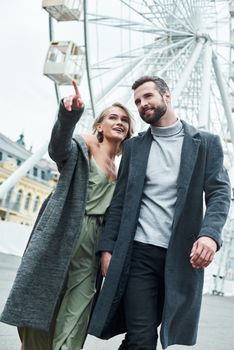 Romantic date outdoors. Young couple walking at entertainment park holding hands talking smiling joyful woman pointing aside at attraction