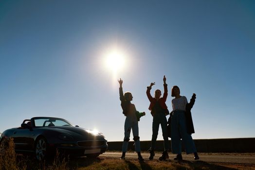 Cheerful young three women are dancing near cabriolet. Beautiful sunset at the background