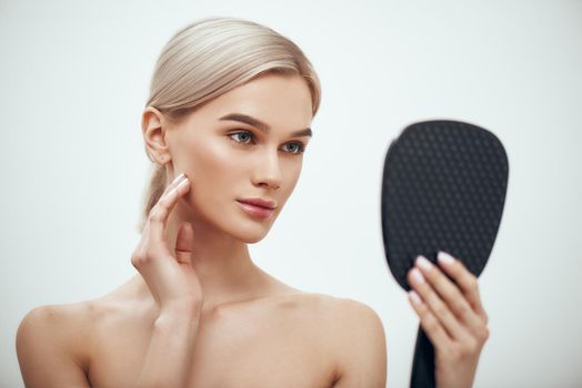 Face care. Portrait of gorgeous blonde woman touching her face and looking in small mirror while standing against grey background. Skin care. Cosmetics concept