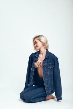 Denim style. Full length of attractive young woman in jeans wear smiling and looking aside while sitting on the floor against grey background in studio. Denim clothes. Fashion concept. Beauty