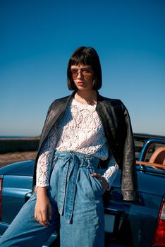 Young woman near roofless car and smiling outdoors on sunny day near the empty road. No people, sportive blue car