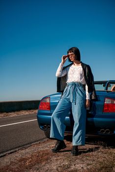 Young attractive stylish woman near roofless car wearing glasses and smiling outdoors on sunny day near the empty road. No people, sportive blue car