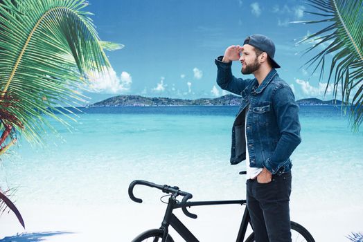 Beach vacation. Young stylish man standing on the beach with bicycle looking forward excited