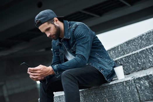 Outdoors leisure. Young stylish man sitting on stairs on city street with cup of hot coffee using application on smartphone joyful