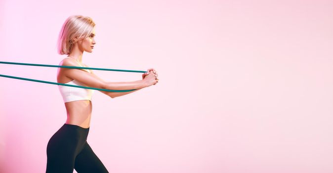 Active lifestyle. Sporty beautiful woman in white top and black leggings exercising with resistance band while standing against pink background in studio. Sport concept. Sport equipment. Web banner