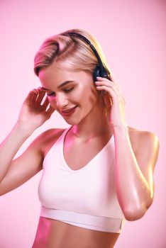 Enjoying music. Close up of cute and sporty woman in headphones listening music and smiling while standing against pink background. Sport. Music concept